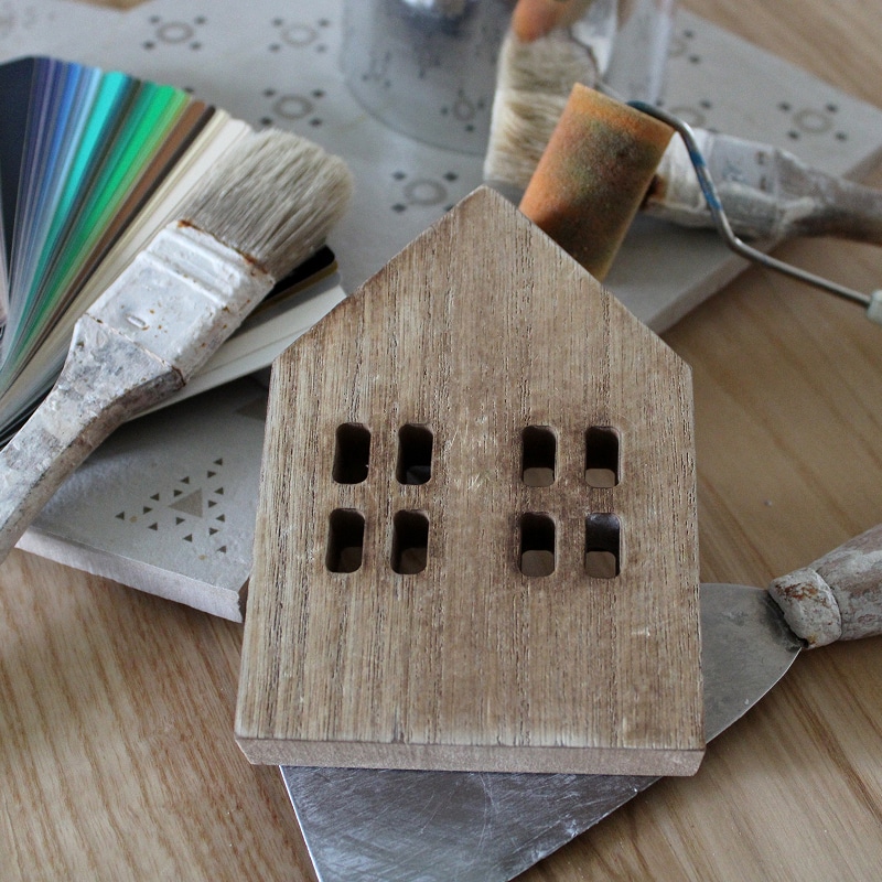 Maquette de maison en bois rustique entourée d'outils de rénovation : nuancier de couleurs, pinceaux sales et spatule, évoquant la décoration intérieure.