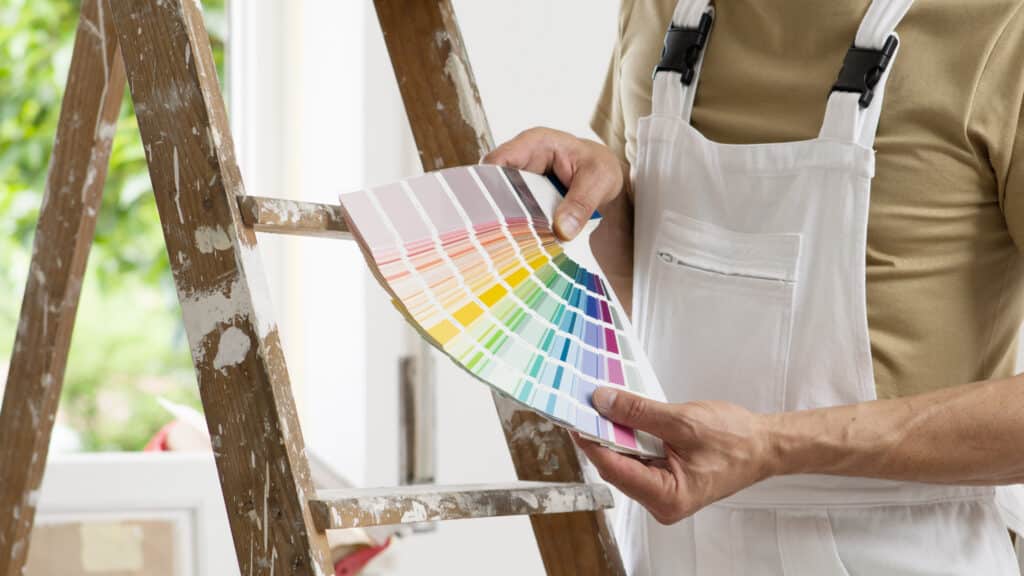 Choix couleurs du peintre : le nuancier idéal pour la rénovation. Peintre en salopette blanche tient un nuancier coloré devant une vieille échelle maculée de peinture, faisant le choix final des teintes.