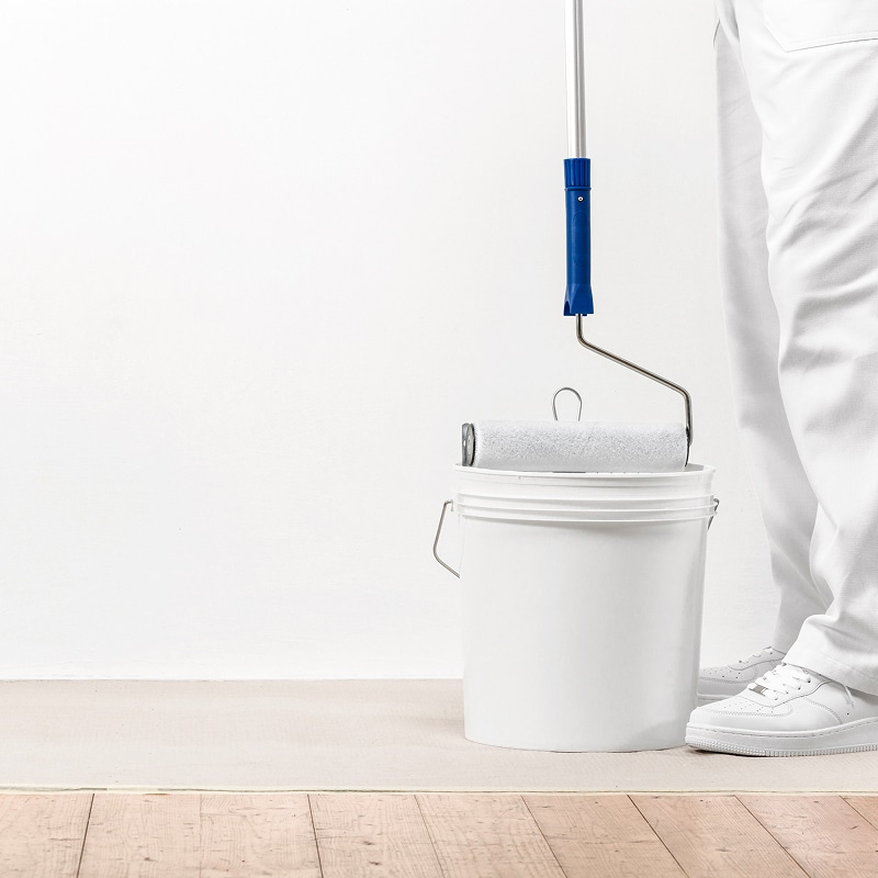 Peintre en tenue blanche et baskets blanches trempe un rouleau à peinture blanc dans un seau de peinture blanche, prêt à peindre un mur blanc.
